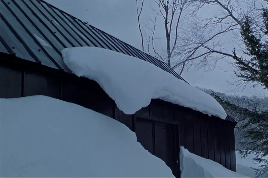 A slab of settled snow releasing off the slope of a dark standing seam metal roof in upstate New York